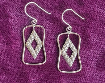 May include: A pair of silver dangle earrings featuring a rectangular wire frame with rounded corners. Inside each frame hangs a diamond-shaped charm with a textured, dotted surface. The earrings are attached to simple silver ear wires for hanging.