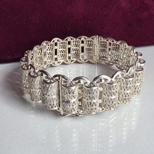 May include: A silver filigree bracelet composed of repeating cylindrical links with intricate wirework patterns. The bracelet features a secure box clasp mechanism and a polished metallic finish, showcasing a detailed openwork design throughout the piece.