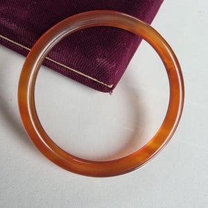 May include: A smooth, circular bangle bracelet crafted from translucent, deep orange-brown material. The bangle features a polished, glossy finish with subtle variations in colour density throughout the ring, creating a warm, glowing appearance.