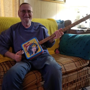 May include: A homemade cigar box guitar featuring a Wonder Woman lunchbox as the body. The guitar has a wooden neck and fretboard, with strings and tuning pegs. The lunchbox is blue, yellow, and red, with a Wonder Woman graphic.