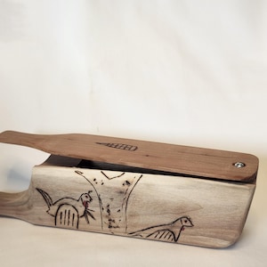 May include: A wooden duck call with a light brown lid and a lighter wood base. The base features etched bird and tree designs. The lid has a small etched design. The call is open, revealing the interior.