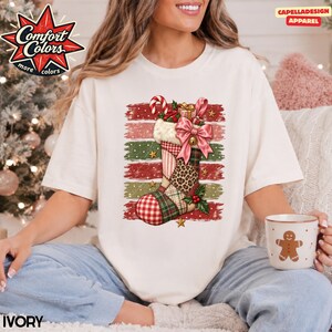May include: An ivory t-shirt featuring a graphic of a Christmas stocking with a patchwork design of plaid, leopard print, and gingham patterns. The stocking is adorned with a large pink bow, a candy cane, and holly, set against a distressed horizontal striped background.