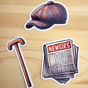 K&ouml;nnte beinhalten: Drei Aufkleber im Vintage-Stil: eine braune und graue Newsboy-M&uuml;tze, ein brauner Gehstock und ein Stapel Zeitungen mit dem in Rot gedruckten Wort "NEWSIES".
