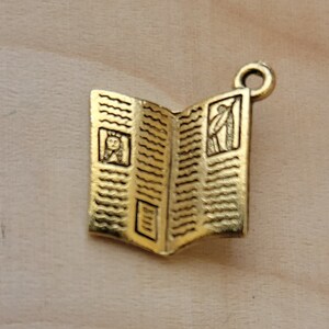 K&ouml;nnte beinhalten: Goldfarbener Metallanh&auml;nger in Form eines ge&ouml;ffneten Buches mit einem detaillierten Design. Das Buch hat einen dekorativen Rand und zwei kleine Bilder im Inneren.
