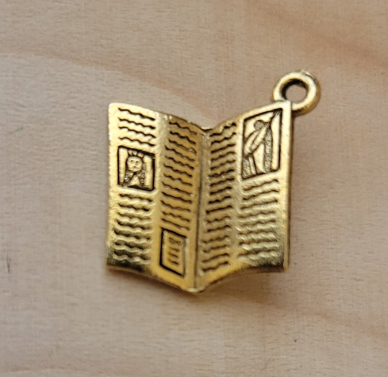 K&ouml;nnte beinhalten: Goldfarbener Metallanh&auml;nger in Form eines ge&ouml;ffneten Buches mit einem detaillierten Design. Das Buch hat einen dekorativen Rand und zwei kleine Bilder im Inneren.