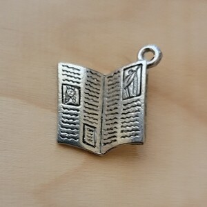 K&ouml;nnte beinhalten: Silberner Anh&auml;nger in Form eines ge&ouml;ffneten Buches mit einem dekorativen Design. Das Buch hat oben eine Schlaufe zum Anbringen an einem Armband oder einer Halskette.