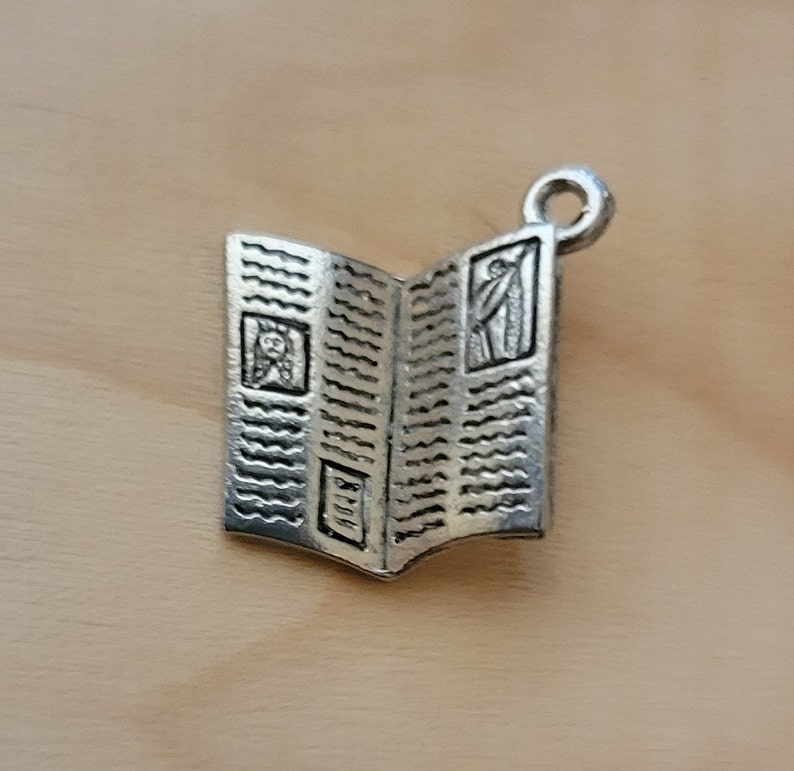 K&ouml;nnte beinhalten: Silberner Anh&auml;nger in Form eines ge&ouml;ffneten Buches mit einem dekorativen Design. Das Buch hat oben eine Schlaufe zum Anbringen an einem Armband oder einer Halskette.