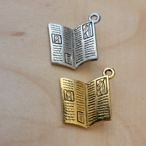 K&ouml;nnte beinhalten: Zwei silberne und goldene Anh&auml;nger in Form von ge&ouml;ffneten B&uuml;chern. Die Anh&auml;nger haben ein detailliertes Design mit einem kleinen Bild auf jeder Seite.