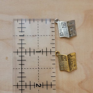 K&ouml;nnte beinhalten: Zwei Anh&auml;nger in Form von Zeitungen, einer in Silber und einer in Gold, mit der Aufschrift "City News" auf der Vorderseite. Die Anh&auml;nger sind neben einem Lineal abgebildet, das etwa 2,5 cm lang ist.
