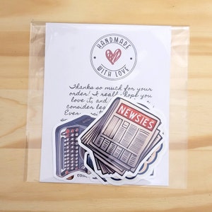 K&ouml;nnte beinhalten: Eine handgeschriebene Notiz auf wei&szlig;em Karton mit einem roten Herzlogo, auf dem "Handmade with Love" steht. Die Notiz lautet: "Vielen Dank f&uuml;r deine Bestellung! Ich hoffe wirklich, dass du sie liebst und eine Bewertung hinterl&auml;sst!" Auf dem Karton befinden sich drei Aufkleber: eine Vintage-Schreibmaschine, ein Stapel Zeitungen mit dem Wort "NEWSIES" oben und ein kleiner runder Aufkleber mit dem Wort "CO" darauf.