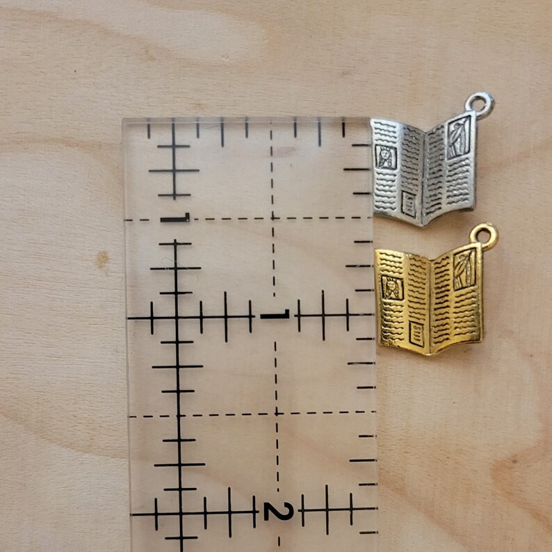 K&ouml;nnte beinhalten: Zwei Buch-Anh&auml;nger, einer in Silber und einer in Gold, mit detailliertem Design, werden neben einem Lineal gezeigt. Das Lineal ist in Zoll eingeteilt.