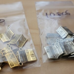 K&ouml;nnte beinhalten: Zwei Beutel mit Metall-Anh&auml;ngern in Form von gefalteten Zeitungen. Der Beutel links enth&auml;lt 10 goldene Anh&auml;nger und der Beutel rechts enth&auml;lt 10 silberne Anh&auml;nger. Die Beutel sind mit "10" und "10 Stk" beschriftet.