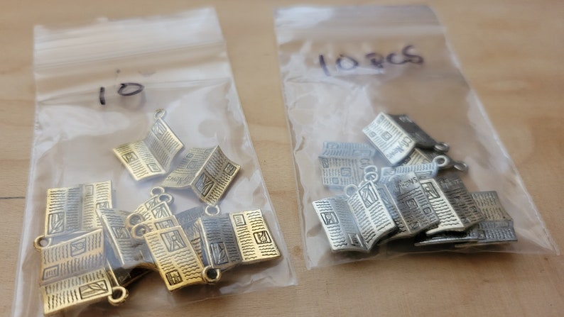 K&ouml;nnte beinhalten: Zwei Beutel mit Metall-Anh&auml;ngern in Form von gefalteten Zeitungen. Der Beutel links enth&auml;lt 10 goldene Anh&auml;nger und der Beutel rechts enth&auml;lt 10 silberne Anh&auml;nger. Die Beutel sind mit "10" und "10 Stk" beschriftet.