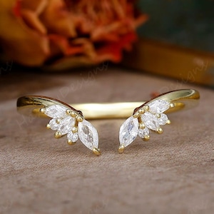 Könnte beinhalten: Ein goldfarbener Ring mit geschwungenem Design, der auf beiden Seiten mit weißen Edelsteinclustern verziert ist. Der Ring hat ein glattes, poliertes Band, und die Edelsteine sind in einer zarten Anordnung angeordnet. Der Ring ist wahrscheinlich ein Schmuckstück.