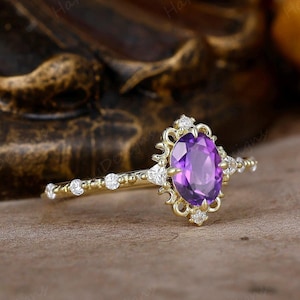 Op de afbeelding: Een delicate gouden ring met een paarse ovale edelsteen omringd door kleinere diamanten. De ring heeft een vintage-geïnspireerd ontwerp met ingewikkelde details.