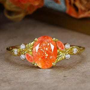 May include: A gold ring featuring an oval, orange gemstone as the centerpiece. The ring is adorned with smaller orange stones and clear accent stones, set within a leaf and vine design.