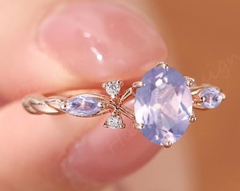 May include: A rose gold ring featuring a large oval-cut, light purple gemstone as the centerpiece. The ring has smaller, light purple stones on either side and a delicate, twisted band. Small clear stones are set around the center stone.