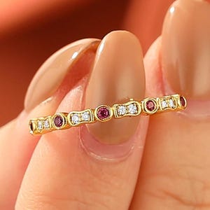 May include: A thin gold band ring featuring an alternating pattern of small round red gemstones and clear white stones set in individual metal bezels. The design consists of a repeating sequence of one red stone followed by two white stones.