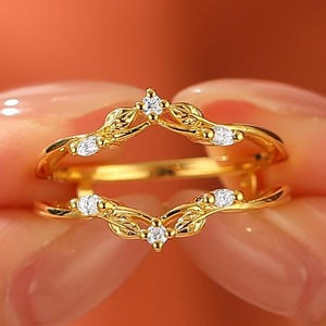 May include: A gold-colored ring with a leaf design and small, clear gemstones. The ring has two bands, with the top band shaped like a crown. The gemstones are evenly spaced along the leaf pattern.