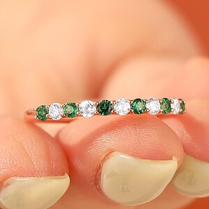 Puede incluir: Un anillo de oro rosa con una fila de piedras preciosas verdes y blancas alternas. El anillo se sostiene en una mano sobre un fondo naranja. Las piedras preciosas son redondas y están espaciadas.