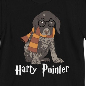 Puede incluir: Camiseta negra con una ilustración de dibujos animados de un cachorro de Braco Alemán de Pelo Corto con una bufanda de Hogwarts y gafas. El texto "Harry Pointer" está impreso debajo de la imagen.