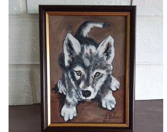 May include: A framed oil painting of a grey and white wolf pup with amber eyes, set against a solid brown background. The artwork is enclosed in a dark brown wooden frame with a thin gold inner border. Two paintbrushes lie in front of the frame.