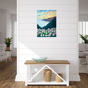 Puede incluir: Una pintura de un paisaje mar&iacute;timo con vista a una costa y un campo de flores rosas en primer plano. El cielo es de un azul vibrante con nubes y el sol se pone en el horizonte.