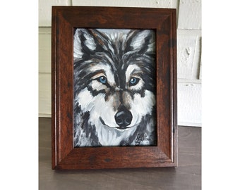 May include: A framed painting of a wolf with blue eyes. The wolf is painted in shades of gray and white, with a dark gray nose and mouth. The painting is set in a dark brown frame.