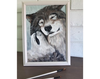 May include: A framed oil painting depicting two wolves in shades of gray, brown, and white. One wolf nuzzles the other, which has its eyes closed. The artwork is enclosed in a light-colored wooden frame, with two paintbrushes resting in front.