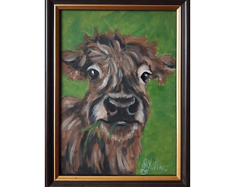 May include: A framed oil painting of a brown Highland cow with shaggy fur, chewing on a blade of grass against a vibrant green background. The artwork is enclosed in a dark brown wooden frame with a thin gold inner border and a signature in the corner.