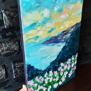 Puede incluir: Una pintura de un paisaje mar&iacute;timo con una cordillera en el fondo. El cielo es de un azul vibrante con nubes blancas y un sol amarillo. El agua es de un azul turquesa y la cordillera es de un verde oscuro. El primer plano es un campo de flores rosas.