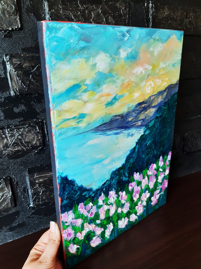 Puede incluir: Una pintura de un paisaje mar&iacute;timo con una cordillera en el fondo. El cielo es de un azul vibrante con nubes blancas y un sol amarillo. El agua es de un azul turquesa y la cordillera es de un verde oscuro. El primer plano es un campo de flores rosas.