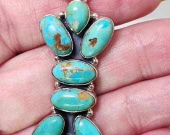 May include: A turquoise pendant with a silver bail. The pendant features oval and teardrop-shaped turquoise stones set in a dark metal frame. The turquoise stones have brown matrix patterns.