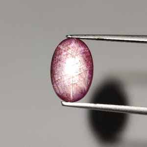 May include: An oval-shaped, polished gemstone held by tweezers. The gem has a pinkish-purple hue with internal light reflections. The surface displays a network of fine, intersecting lines. The gem is set against a neutral gray background.