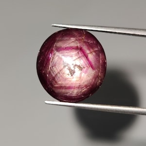 Könnte beinhalten: Ein runder, facettierter Edelstein, der von einer Pinzette gehalten wird. Der Edelstein hat eine tiefrote bis violette Farbe mit inneren Linien und einem leicht durchscheinenden Aussehen. Der Stein ist wahrscheinlich ein Rubin, und die Pinzette ist silberfarben.