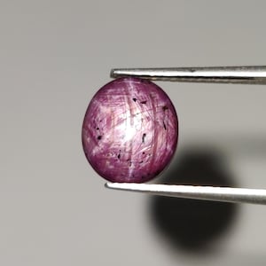 May include: A polished, oval-shaped ruby gemstone, held by tweezers. The gem is a deep magenta color with internal striations and dark inclusions. The light reflects off the surface, creating a star-like effect.