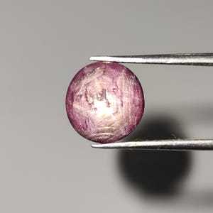 Puede incluir: Una gema redonda y pulida sujeta con pinzas. La gema es de color rosa intenso con una superficie arremolinada y texturizada y un área más clara, casi blanca, en el centro. El fondo es gris neutro.