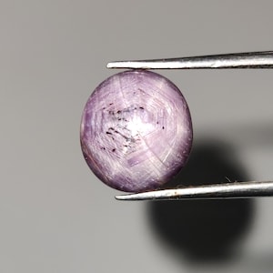 Op de afbeelding: Een ovale, lichtpaarse edelsteen, vastgehouden door een pincet. De edelsteen heeft een stervormig patroon en een enigszins doorschijnend uiterlijk. De kleur is zacht lavendel met donkere insluitsels.
