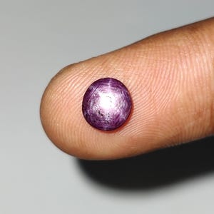 May include: A small, round, purplish-red gemstone with a star-like effect. The gem is held on a fingertip, showcasing its polished surface and internal light play. The stone's color ranges from deep red to violet, with a prominent star pattern.