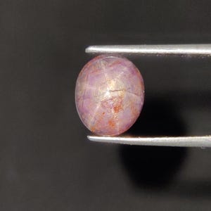 Peut inclure: Une pierre précieuse rubis étoilé de taille cabochon ronde, présentant une teinte rose violacé translucide. La pierre affiche un effet d'étoile à six branches distinct sur sa surface, formé par des lignes lumineuses qui se croisent.