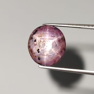 May include: A round, polished gemstone with a purplish-pink hue, held by a pair of silver tweezers. The gem has a smooth surface with internal light reflections and dark inclusions. The background is a neutral gray.