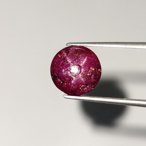 Puede incluir: Una piedra preciosa ovalada de color rojo oscuro con un patrón en forma de estrella en el centro. La piedra preciosa está sujeta por unas pinzas sobre un fondo blanco.