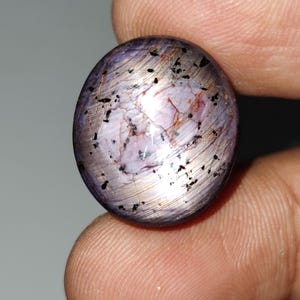 May include: A purple and silver gemstone with black flecks. The gemstone is oval shaped and has a smooth, polished surface.