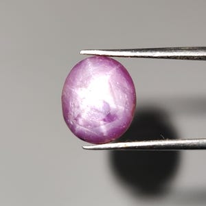 Może przedstawiać: Wypolerowany, owalny, fioletowo-różowy kamień szlachetny trzymany pęsetą. Kamień ma gładką powierzchnię z subtelnym, gwiaździstym efektem. Tło jest neutralnie szare, podkreślające kolor i kształt kamienia.