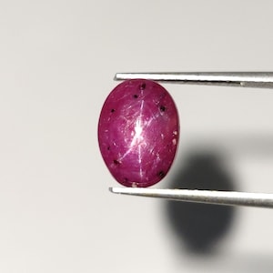Könnte beinhalten: Ein ovaler rosa Sternrubin mit einem sechszackigen Stern in der Mitte. Der Rubin wird von einer Pinzette gehalten.