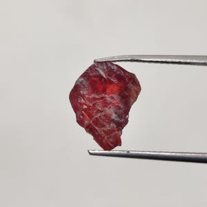May include: A rough-cut, deep red gemstone is held by a pair of silver tweezers against a white background. The gemstone has a textured surface and irregular shape, showcasing its natural form. The color is a rich, saturated red.