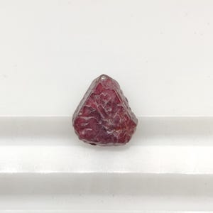 Könnte beinhalten: Ein kleiner, rauer, tiefroter Edelstein in dreieckiger Form mit einer texturierten, facettierten Oberfläche. Der Stein zeigt verschiedene Schattierungen von Karmesinrot und Kastanienbraun mit natürlichen Vertiefungen und unregelmäßigen Kanten.