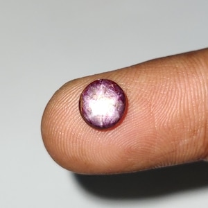 May include: A round, polished gemstone with a deep purple hue and a star-like effect. The gem is held on a fingertip, showcasing its size and color. The stone's surface reflects light, creating a shimmering appearance. This gemstone could be used in jewelry.