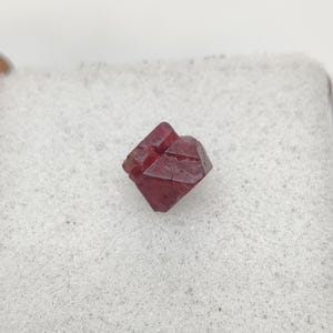 Peut inclure: Un petit cristal rouge foncé de forme naturelle, présentant une géométrie cubique ou octaédrique distincte. Ce spécimen minéral possède des arêtes vives et anguleuses ainsi qu'une texture de surface légèrement translucide et facettée.