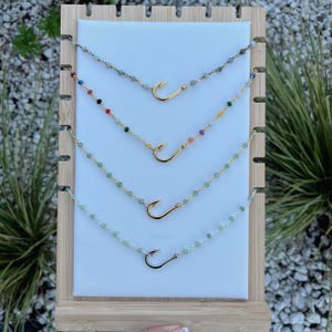 May include: Four gold-colored necklaces with fish hook pendants. Each necklace features a different colored bead strand. The necklaces are displayed on a white background with a wooden frame, showcasing the jewelry's design and colors.
